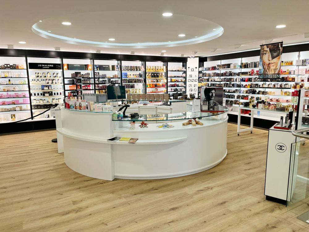 Innenansicht einer großzügigen Parfümerie mit hellen Räumen, Holzfußboden und einem runden weißen Verkaufstresen in der Mitte. Im Hintergrund stehen beleuchtete Regale mit Parfums und Pflegeprodukten verschiedener Luxusmarken wie Jo Malone London, Tom Ford und Chanel. Die Atmosphäre wirkt modern, elegant und einladend.