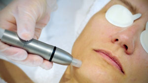 Kosmetische Gesichtsbehandlung: Eine Frau liegt entspannt mit geschützten Augen, während ein kosmetisches Handgerät von einer behandschuhten Hand an der Wangenpartie angewendet wird