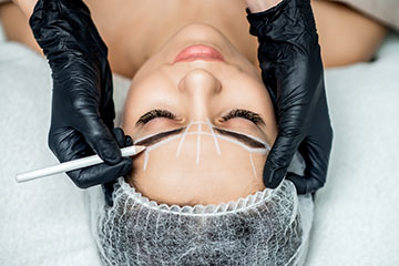 Frau liegt mit geschlossenen Augen auf einer Liege, während eine Kosmetikerin mit schwarzen Handschuhen die Augenbrauen für ein Microblading mit weißen Linien vermisst und markiert.