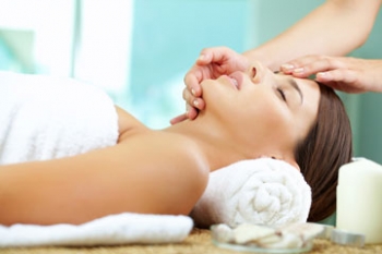 Frau liegt mit geschlossenen Augen und Handtuch unter dem Kopf auf einer Liege, während eine Kosmetikerin eine Gesichtsbehandlung durchführt.