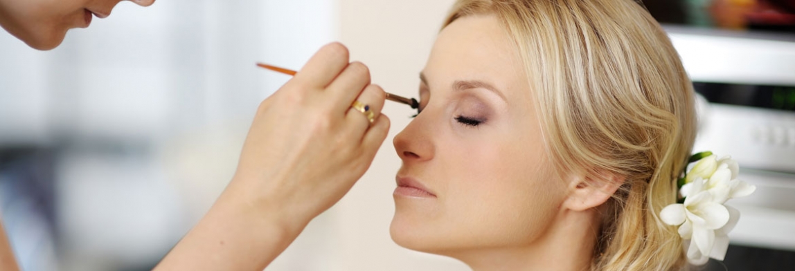 Professionelles Make-up: Eine Visagistin trägt mit einem Pinsel Lidschatten auf das geschlossene Auge einer Frau auf, die entspannt sitzt