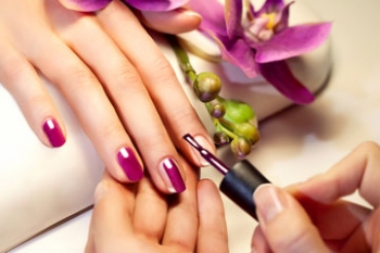 Eine Person trägt pinkfarbenen Nagellack auf die Fingernägel einer anderen Person auf, im Hintergrund eine violette Blüte.