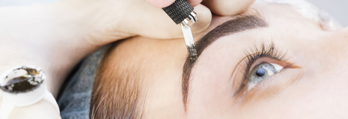 Nahaufnahme einer Microblading-Behandlung: Eine behandschuhte Hand arbeitet mit einem feinen Klingenwerkzeug an der Augenbraue einer Frau, um pigmentierte Härchen zu zeichnen