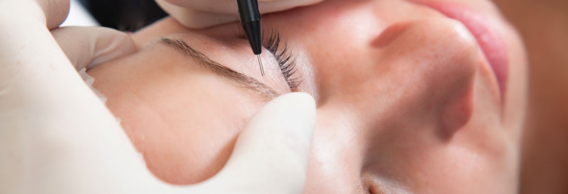 Nahaufnahme einer Permanent-Make-up-Behandlung am Auge: Eine behandschuhte Hand führt ein feines Pigmentiergerät entlang des Wimpernkranzes einer liegenden Frau