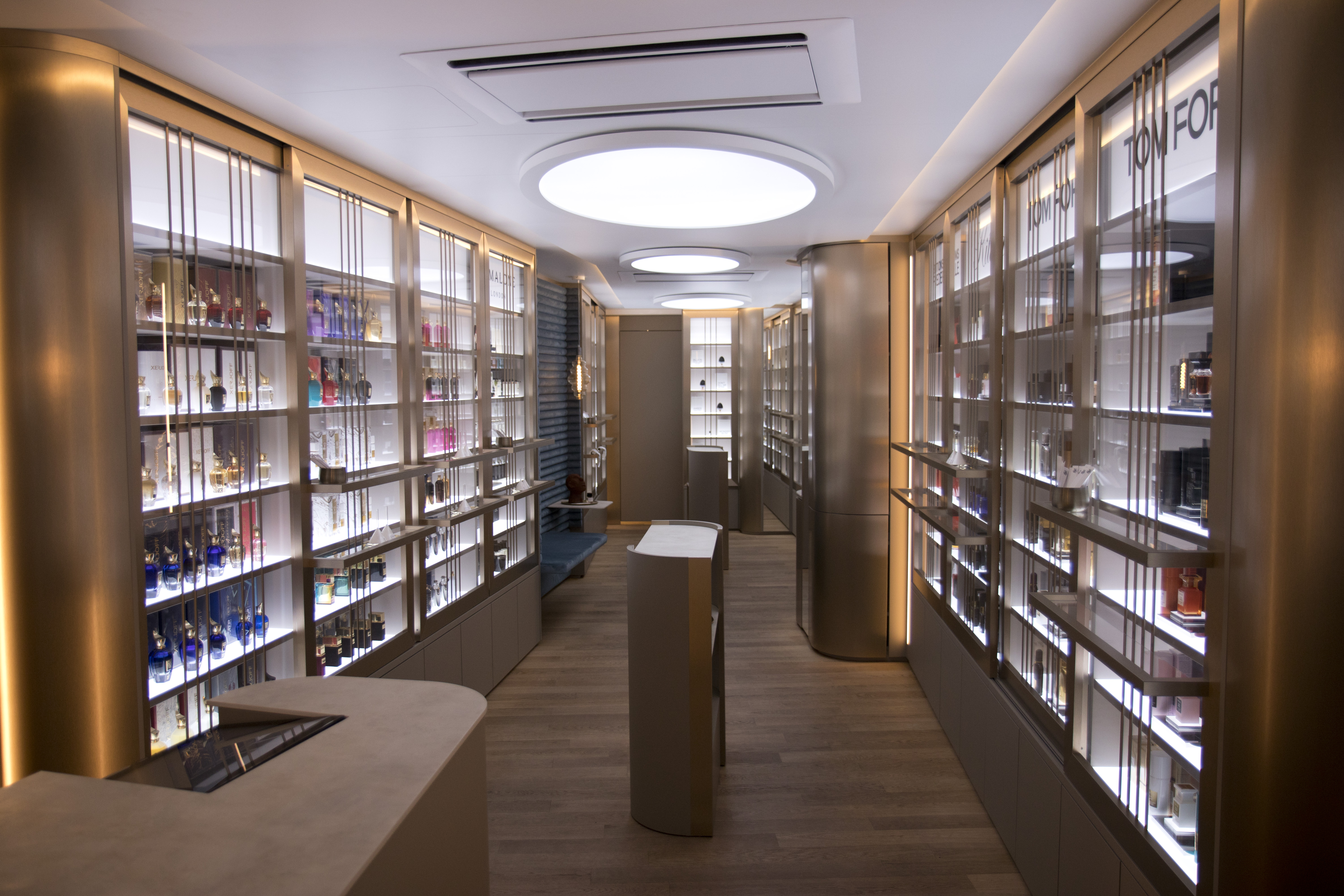 Innenansicht einer modernen Parfümerie mit beidseitig beleuchteten Regalwänden, gefüllt mit hochwertigen Parfums in eleganten Flakons. Die goldfarbenen Rahmen, das warme Licht und die runden Deckenleuchten schaffen eine luxuriöse, einladende Atmosphäre. In der Mitte stehen helle Präsentationstische auf Holzfußboden.