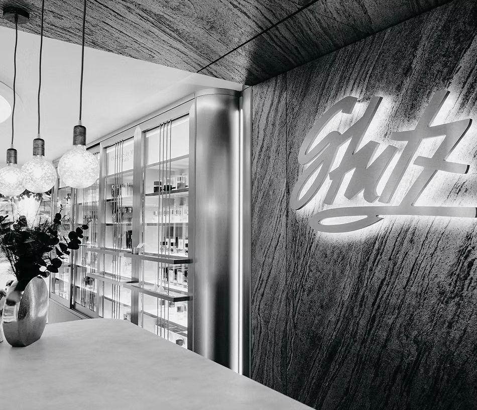 Modern gestalteter Innenraum einer Parfümerie mit beleuchtetem Regal voller Parfümflakons, eleganten Hängelampen und beleuchtetem Schriftzug ‚Schutz‘ an der Wand.